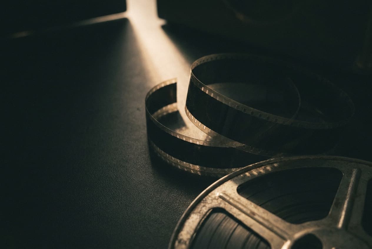 A length of 16mm film unspooled beside a metal reel on a dark surface, lit by a single beam of light.